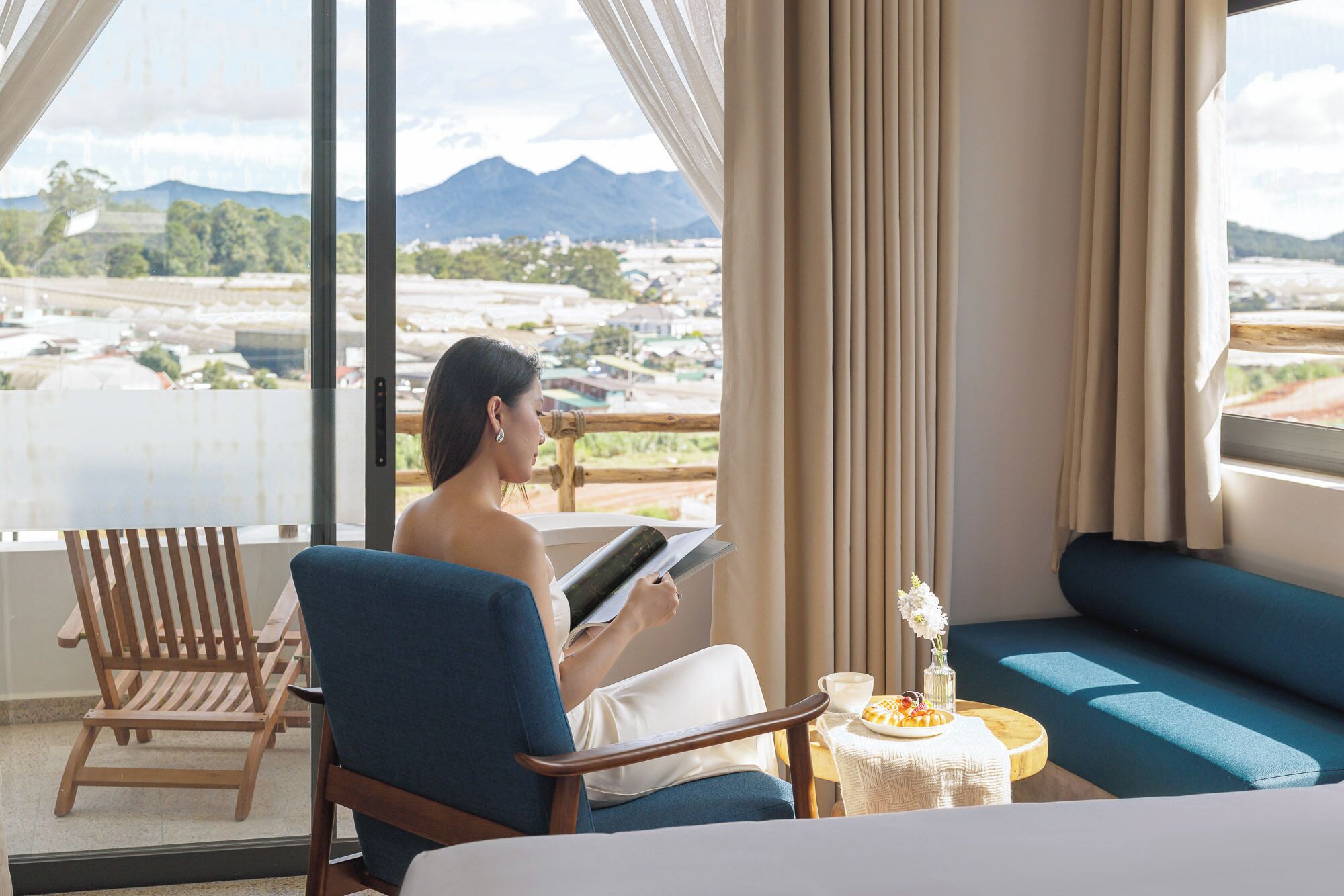 Ein Zimmer mit großem Fenster, einem Bett, einem Sessel und einem kleinen Tisch mit Tasse und Blumen.