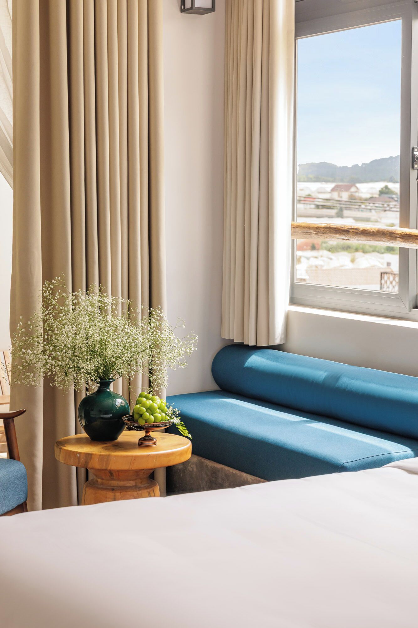 Una habitación con una cama, un sofá azul, un ventanal con cortinas y una mesita con un jarrón de flores.