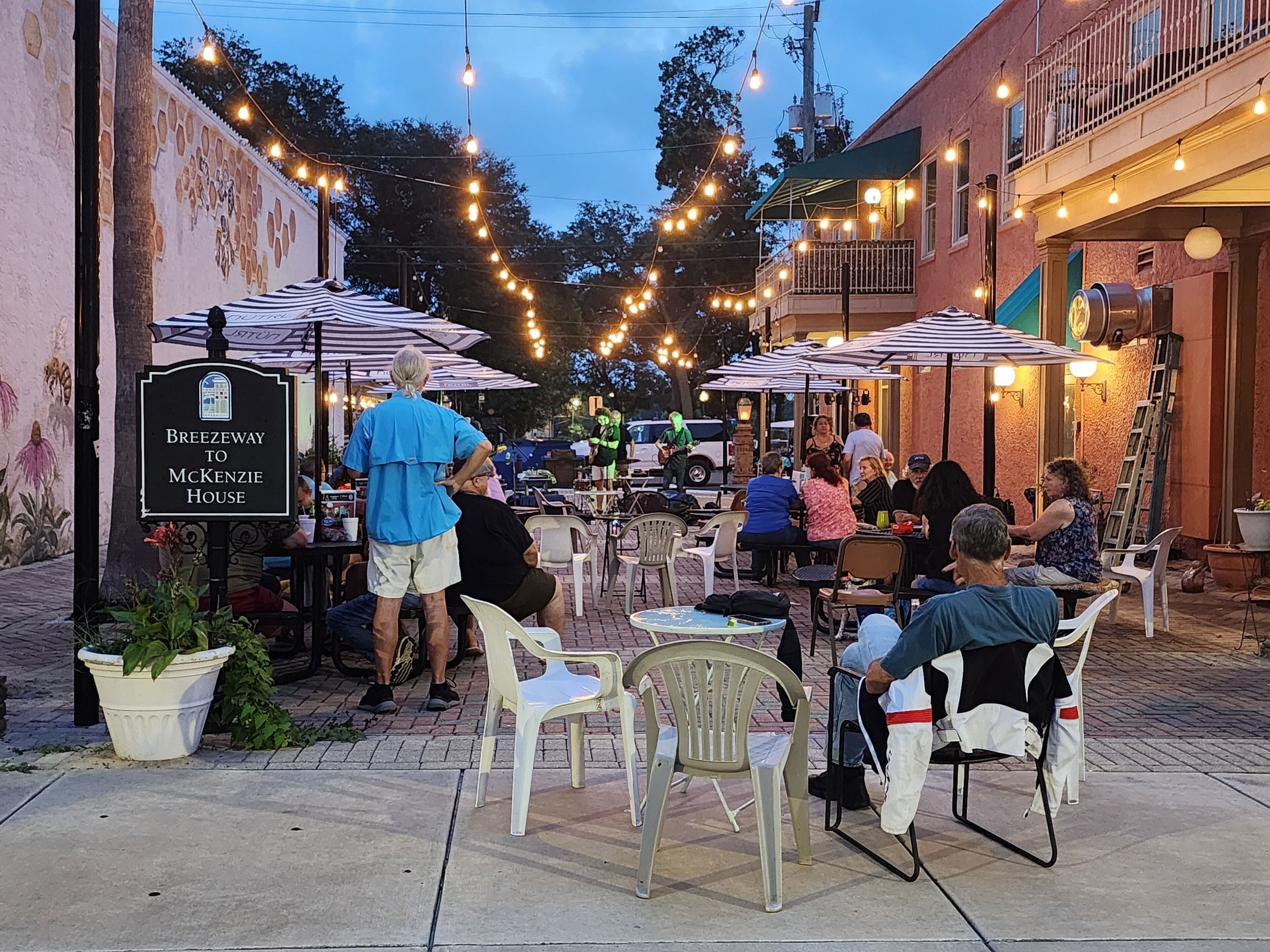 An outdoor seating area with tables and chairs, string lights overhead, and a sign for 'Breezeway to McKenzie House.'