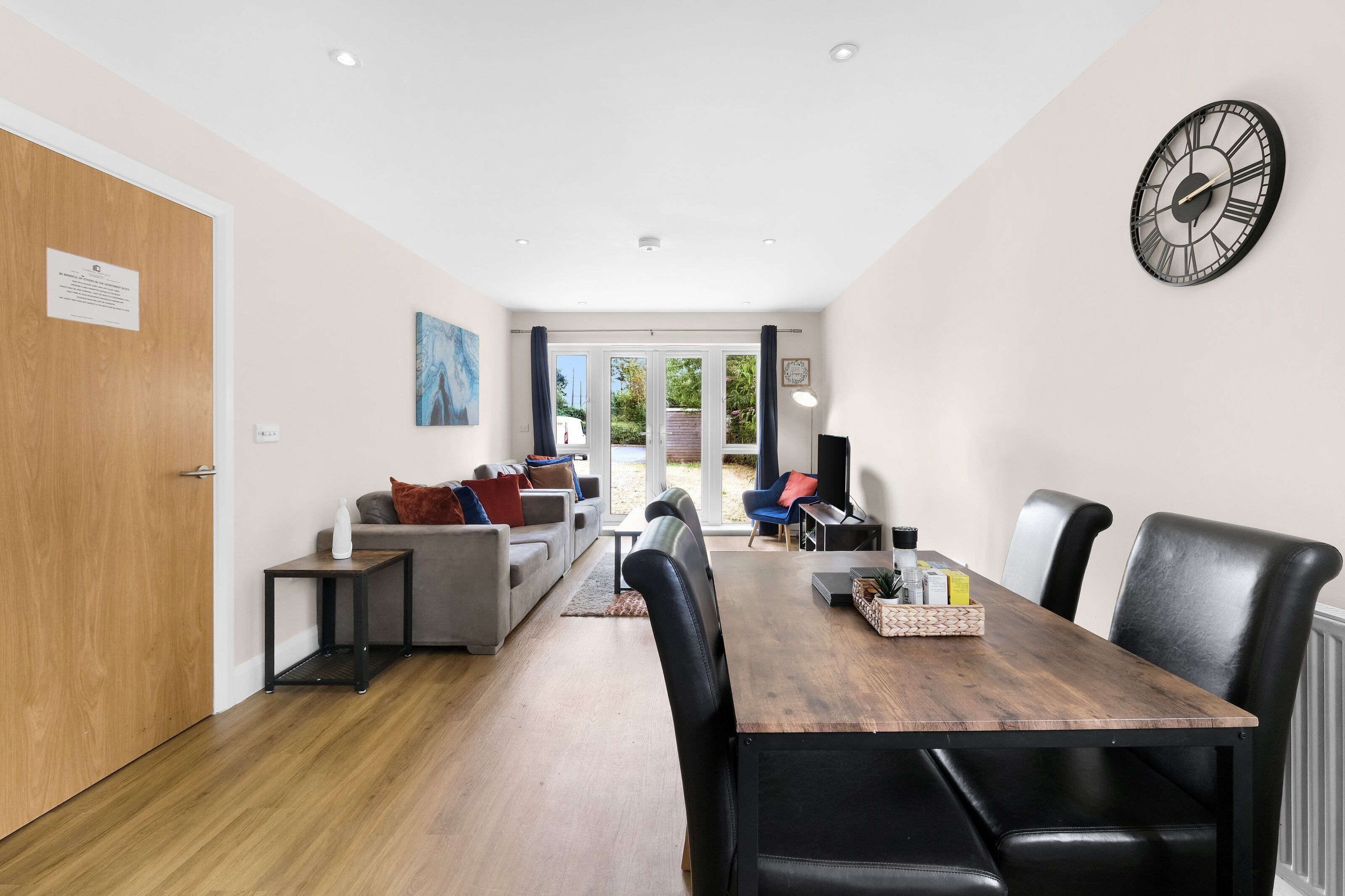 Een moderne woonkamer met een houten eettafel, zwarte lederen stoelen, een grijze bank en een grote wandklok.