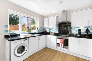 A modern kitchen with a washing machine, stove, oven, and a window with a view of greenery. - Heathrow Living 2BDR 2BA Apt 09-360stays (Stanwell)