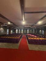 Un salón de conferencias con filas de sillas, alfombra roja y un techo alto con luminarias.