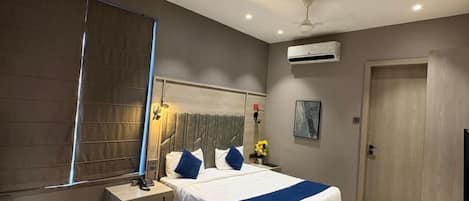 A modern hotel room with a bed, bedside table, chair, desk, and an air conditioning unit.
