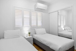 A minimalist bedroom with two beds, a bedside table with a lamp, and a large mirror. - Poolside 2-bed House in Coolum Beach (Sunshine Coast)