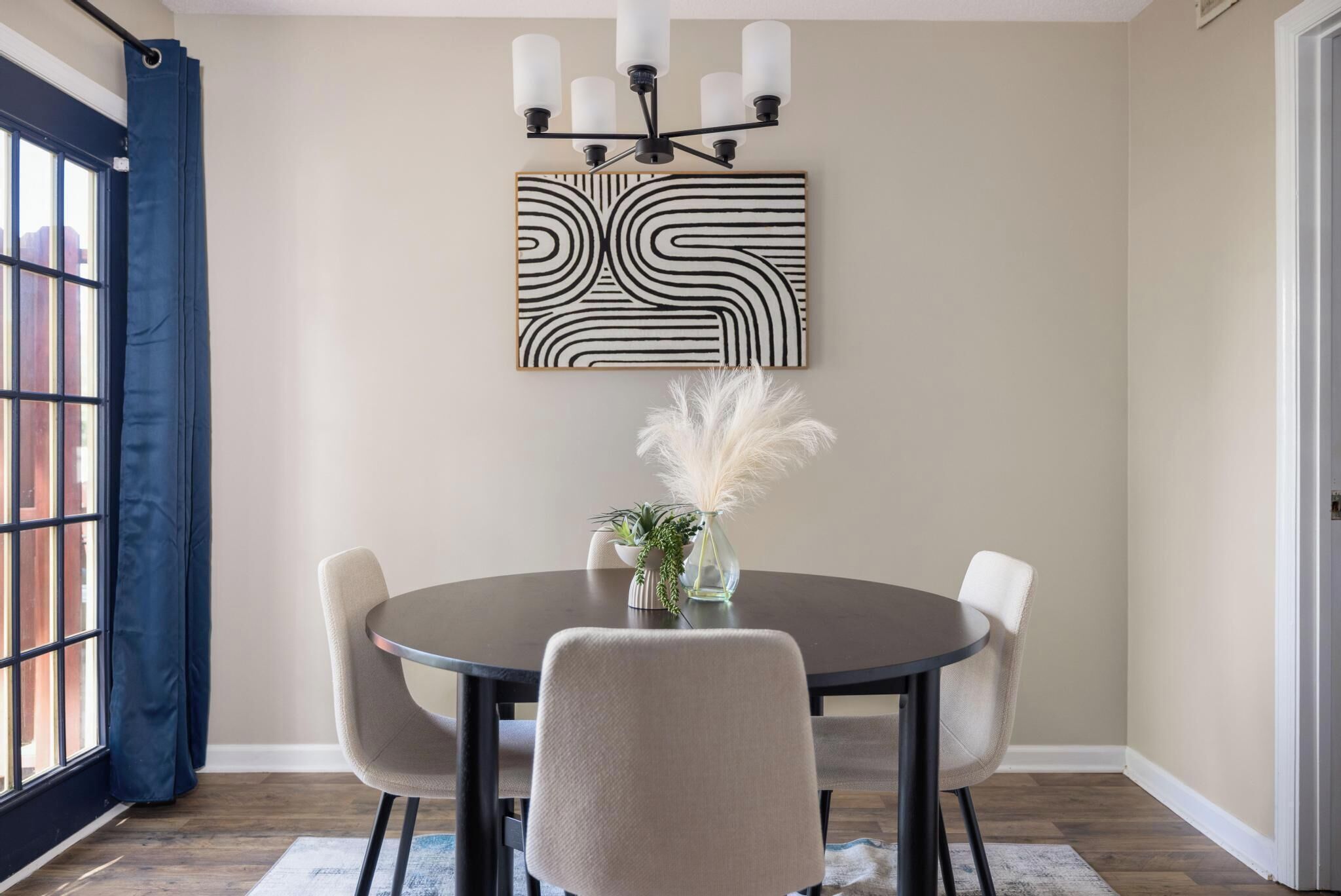 Un coin repas comprenant une table ronde, quatre chaises, un lustre, une œuvre d’art murale et une fenêtre avec des rideaux.