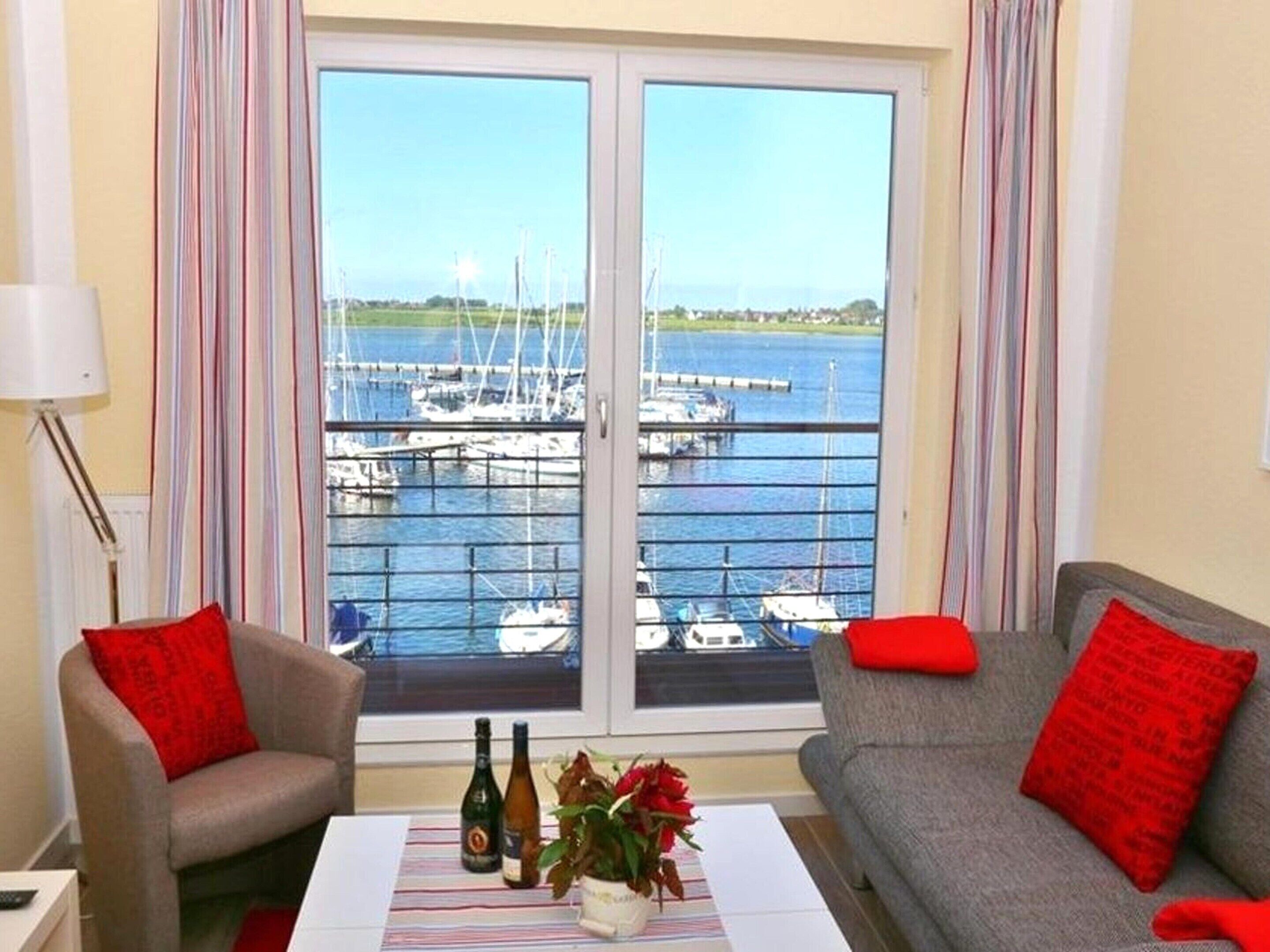 A room with a view of a marina, a sofa, a chair, a table with bottles and flowers, and curtains.