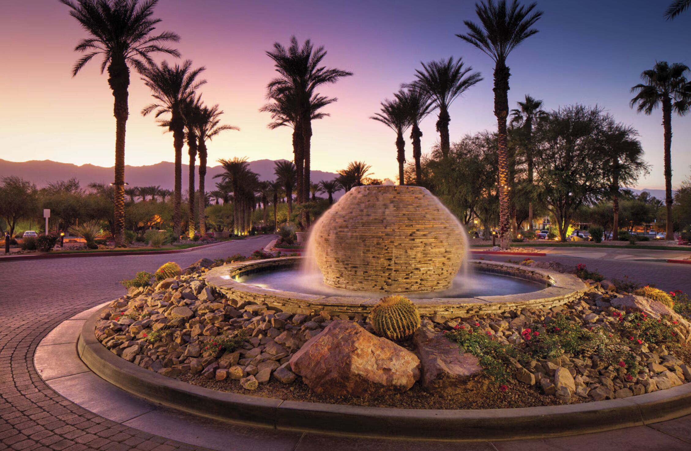 A landscaped fountain with a stone structure, surrounded by rocks and cacti, set against a backdrop of palm trees and mountains.