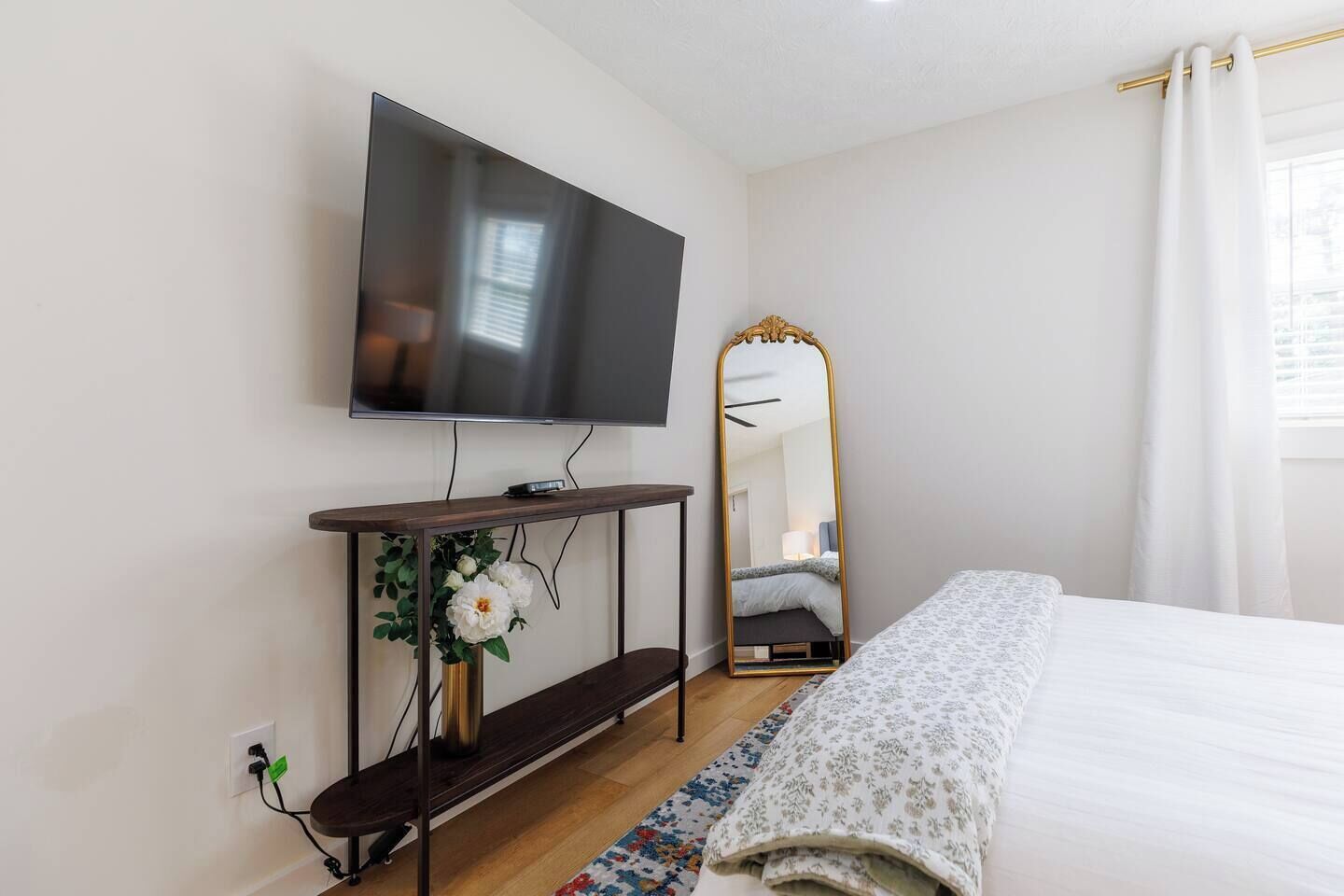 A bedroom with a bed, a TV mounted on the wall, a bedside table with a mirror, and a floral arrangement.