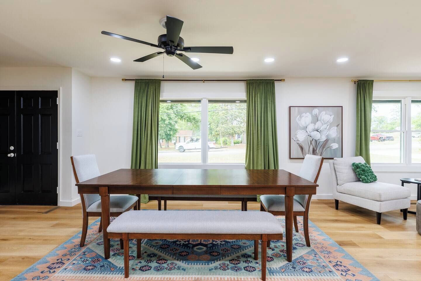 A dining area with a wooden table, chairs, a bench, a ceiling fan, and a view through large windows with green curtains.