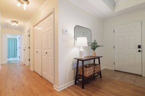 A hallway with wooden flooring, white doors, a wall-mounted mirror, a table with a lamp, and a potted plant.
