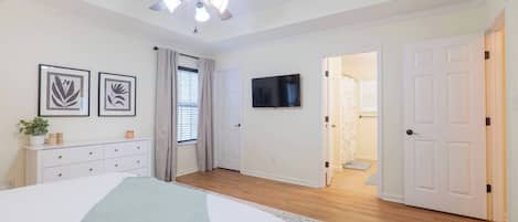 Une chambre à coucher comprenant un lit, un ventilateur de plafond, une télévision, une commode et une porte donnant sur une autre pièce.