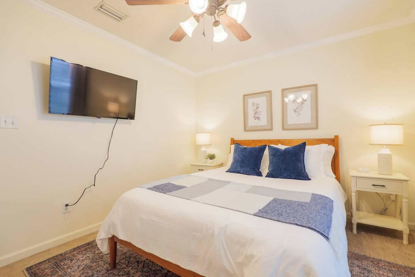 A hotel room with a bed, a ceiling fan, a flat-screen TV mounted on the wall, two bedside lamps, and framed artwork on the wall.