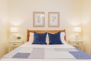 Una cama con cabecera de madera, dos almohadas, una colcha blanca y dos mesitas de noche con lámparas y una planta pequeña.