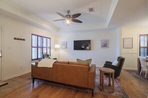 A living room with a brown sofa, a ceiling fan, a flat-screen TV, and a dining area with a table and chairs.