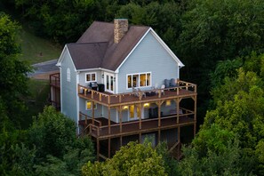 A two-story house with a deck and a chimney, surrounded by dense forest.