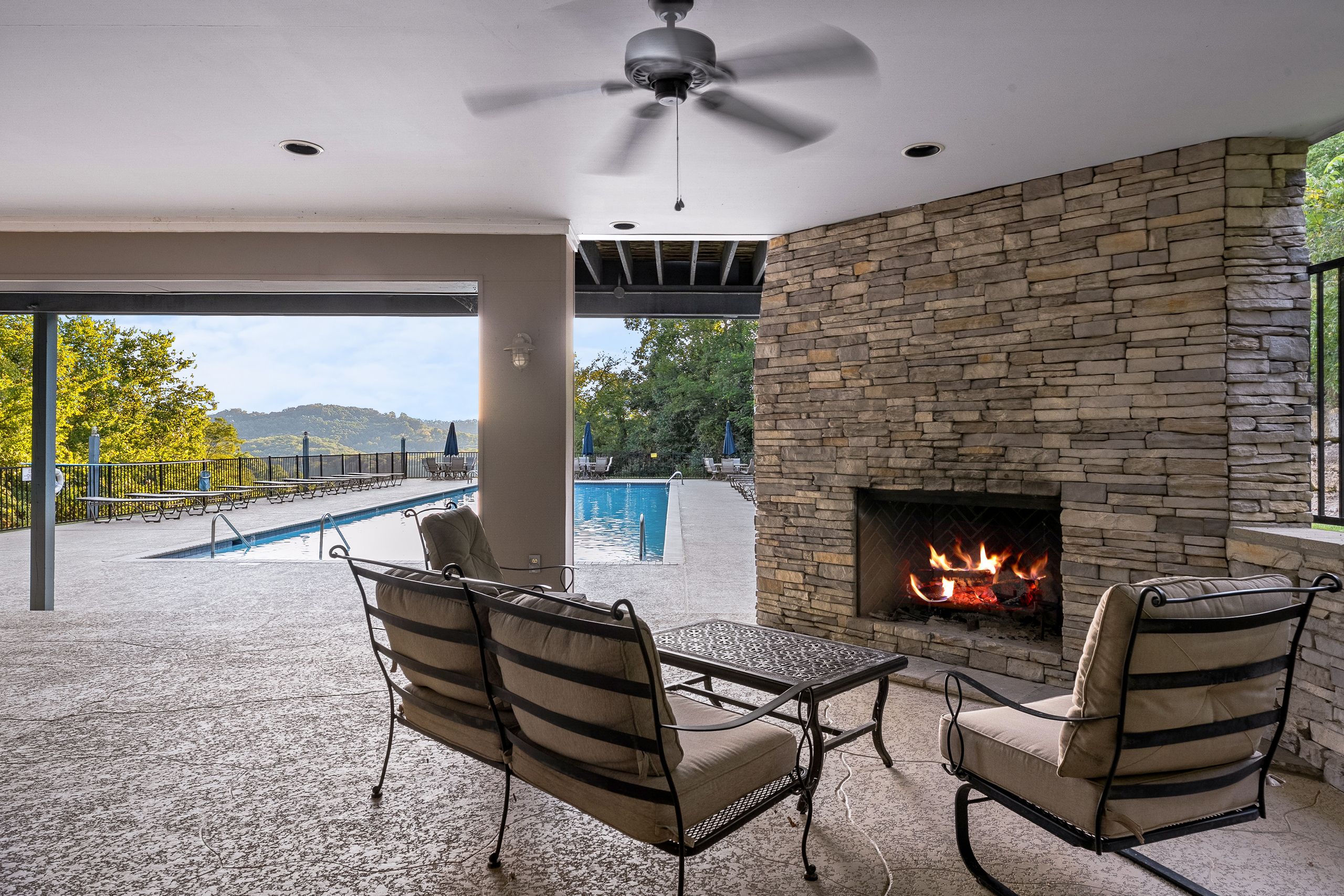 Un patio exterior con chimenea de piedra, dos sillones con cojines, una piscina y vistas a árboles y colinas.