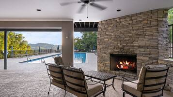 Un patio exterior con chimenea de piedra, dos sillones con cojines, una piscina y vistas a árboles y colinas.