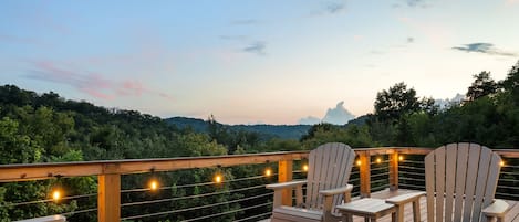 A deck with Adirondack chairs and a built-in fire feature, overlooking a forested landscape.