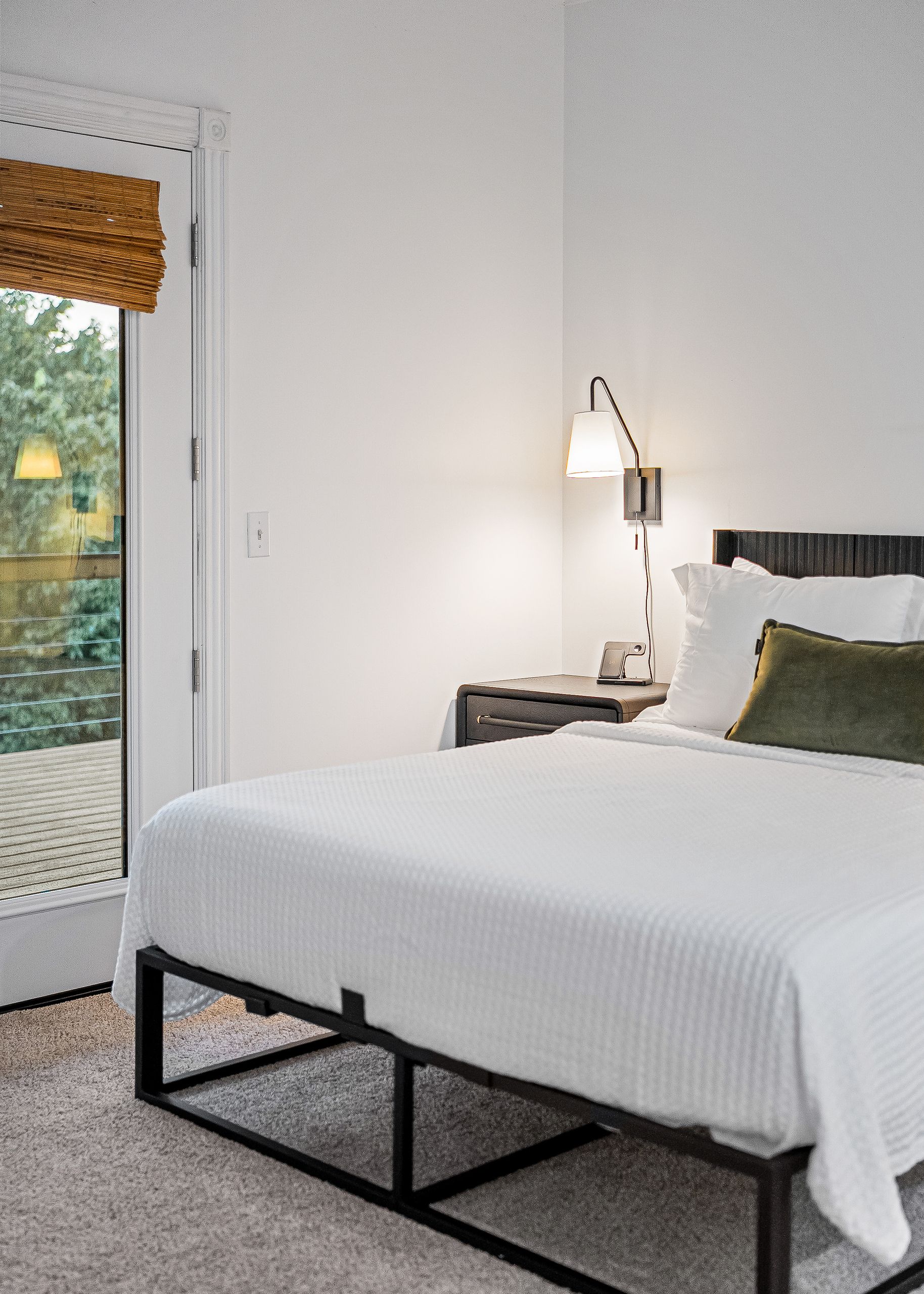 Une chambre d’hôtel avec un lit, une table de chevet, une lampe et une fenêtre donnant sur l’extérieur, équipée de stores.