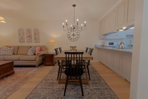 Een woonkamer met een bank, een eettafel met stoelen, een kroonluchter, en een keuken met kasten en apparatuur.