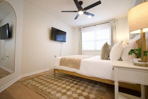 Een slaapkamer met een bed, een plafondventilator, een televisie aan de muur en een nachtkastje met een lamp.