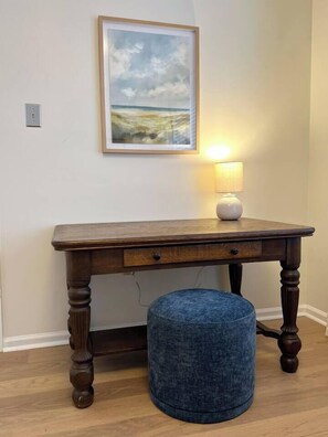 Een houten tafel met een lade, een rond hockertje, een lamp en een lijst met een foto aan de muur.