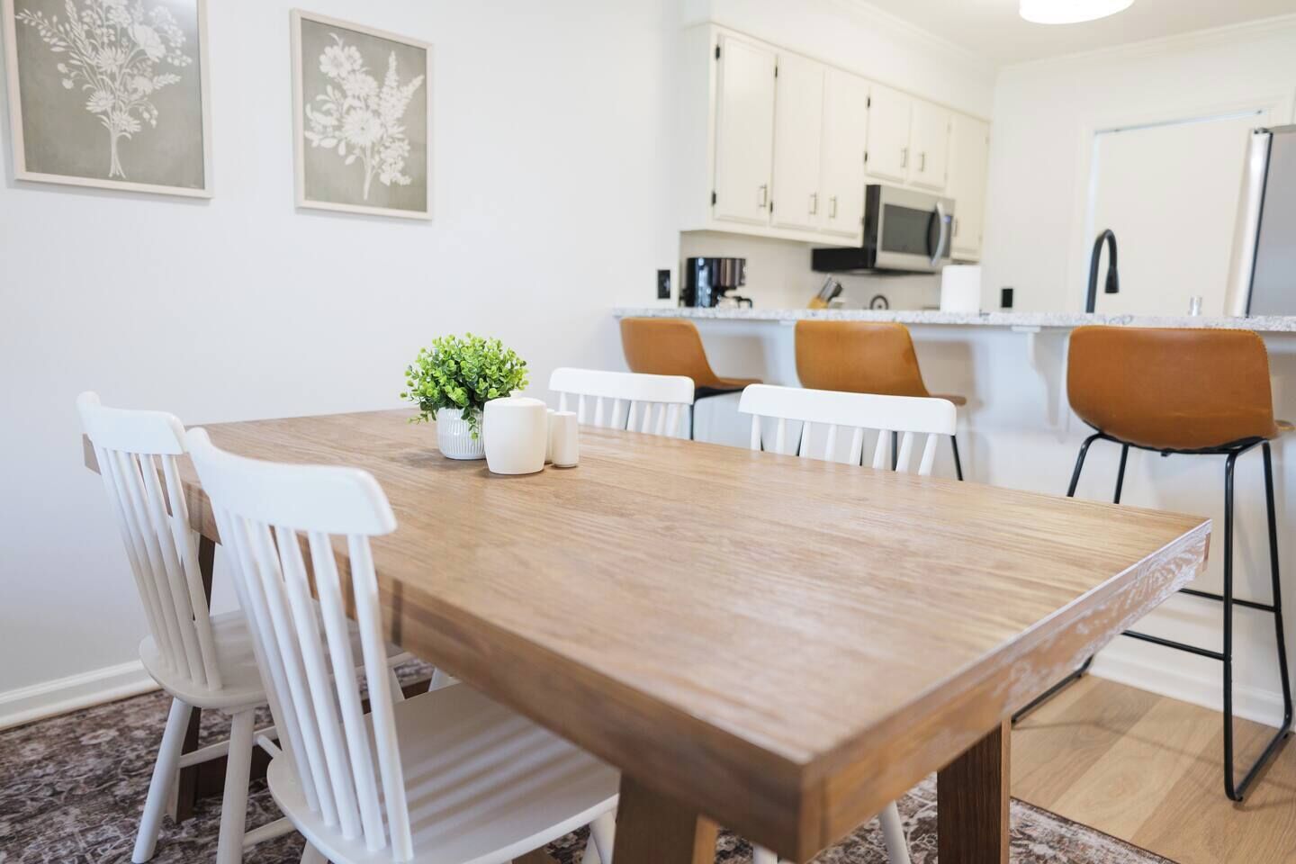 A dining area with a wooden table, white chairs, a small plant, and a kitchen in the background.