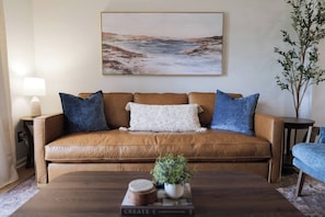 Ein braunes Ledersofa mit blau-weißen Kissen, ein Couchtisch mit Pflanze und Büchern, und ein Landschaftsbild an der Wand.