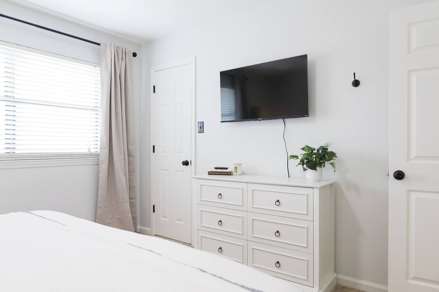 Un dormitorio con cama, cómoda, televisión en la pared y ventana con cortinas.