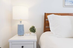 Uma cama bem arrumada com travesseiro branco e cabeceira de madeira, uma mesa de cabeceira branca com abajur, uma pequena planta em vaso e um despertador branco.