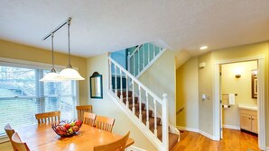 A dining area with a wooden table, chairs, and a fruit bowl. A staircase with white railings leads to another room. - 3 Bedroom Condo with stunning sunset views located in The Homestead Resort! (Glen Arbor)