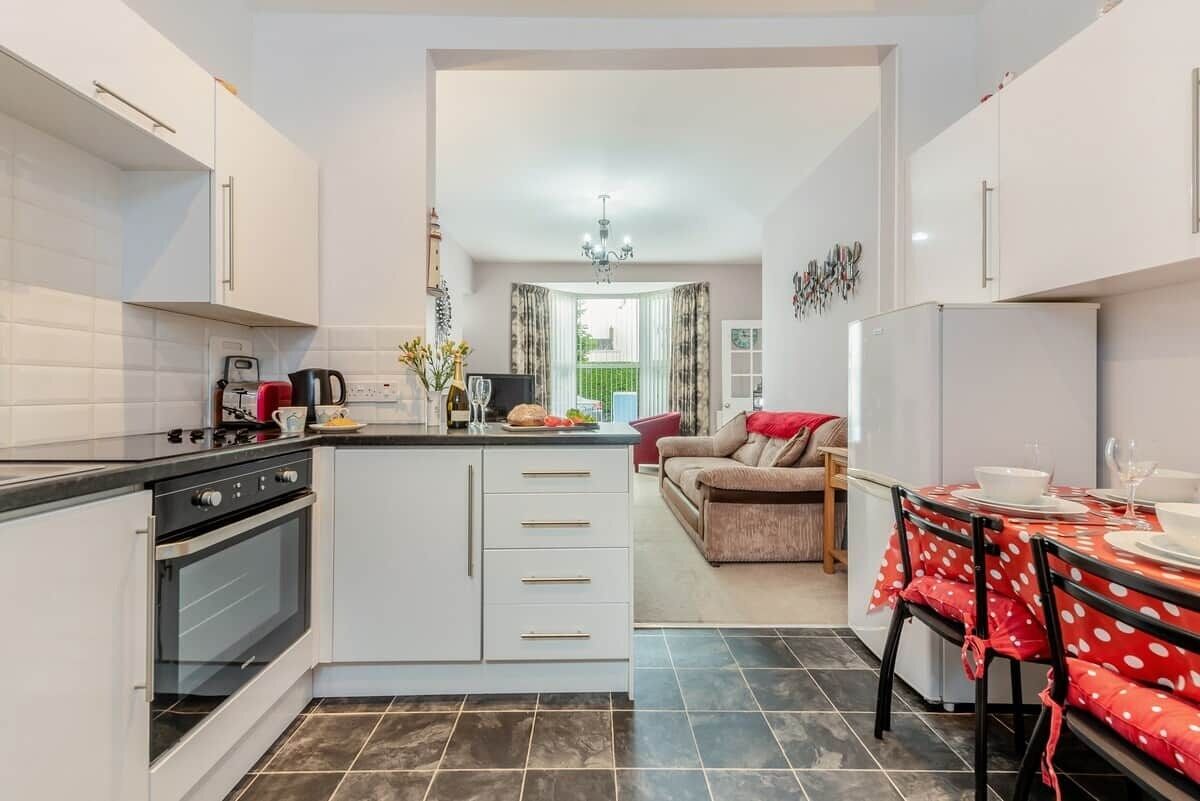 A compact kitchen with a built-in oven, a dining area with a red and white tablecloth, and a living room with a sofa and a red throw blanket.