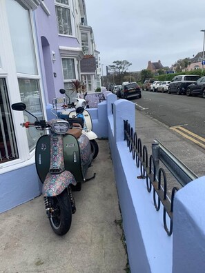 Un trottoir étroit, bordé de scooters et de voitures garées, un mur bleu avec une rambarde métallique, et un immeuble d’habitation avec une fenêtre blanche.