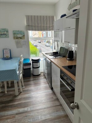 A compact kitchen with a view of the street, a dining area with a table and chairs, and a recycling bin. - Cheriton View | Close to Beach | Sea Glimpse (Tenby)