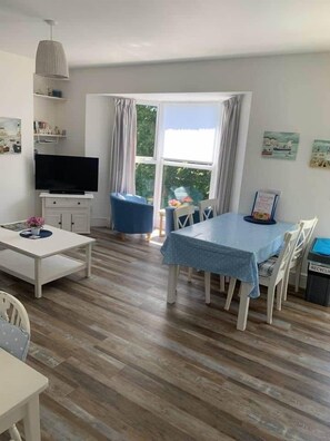Ein Wohnzimmer mit Fernseher, Esstisch, Stühlen und einem fenster mit Vorhängen.