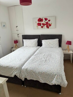 Ein Doppelbett mit weißem Überwurf, zwei Nachttische mit Lampen, ein Kleiderschrank und ein Bild von roten Blumen an der Wand.