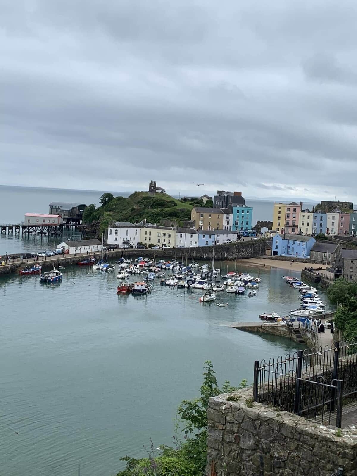 A coastal town with a harbor filled with boats, colorful buildings, and a prominent hill with a structure on top.