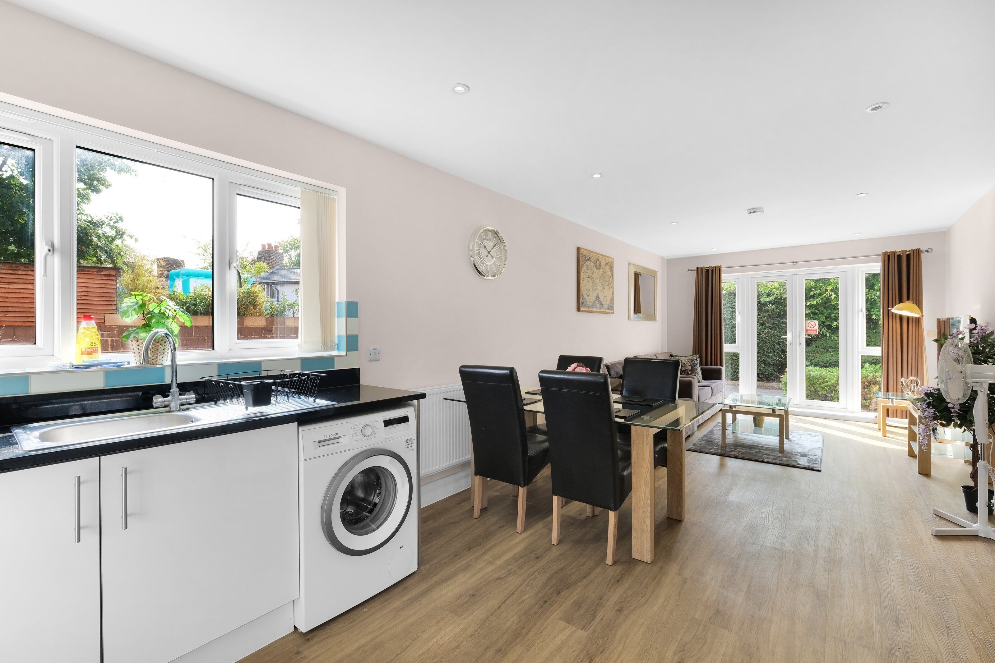 A modern kitchen with a Bosch washing machine, a dining area with a glass table, and a view of the garden through the windows.