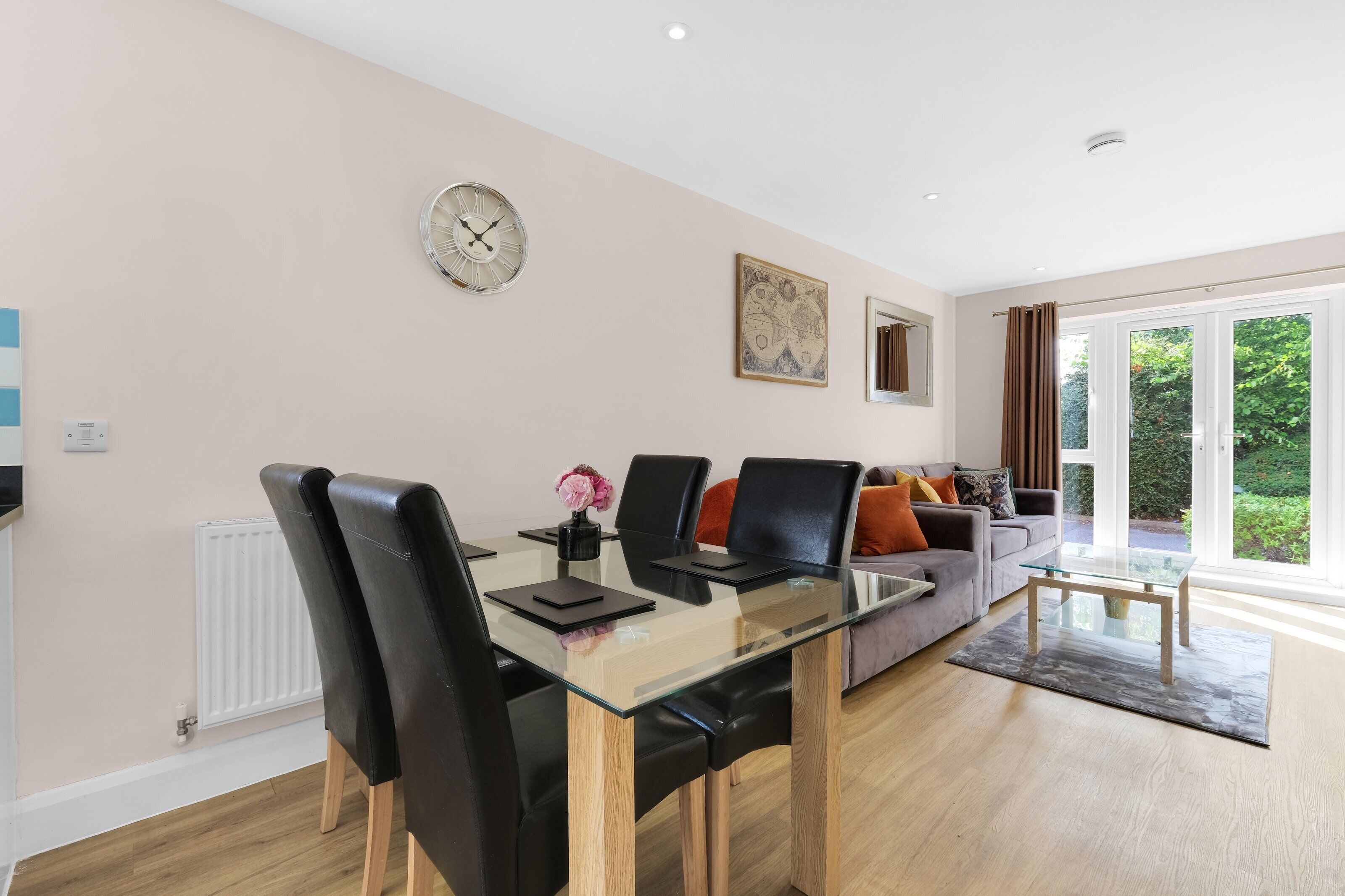 Een moderne woonkamer met een glazen eettafel, zwarte leren stoelen, een bank met kleurrijke kussens, een wandklok en een groot raam met gordijnen.