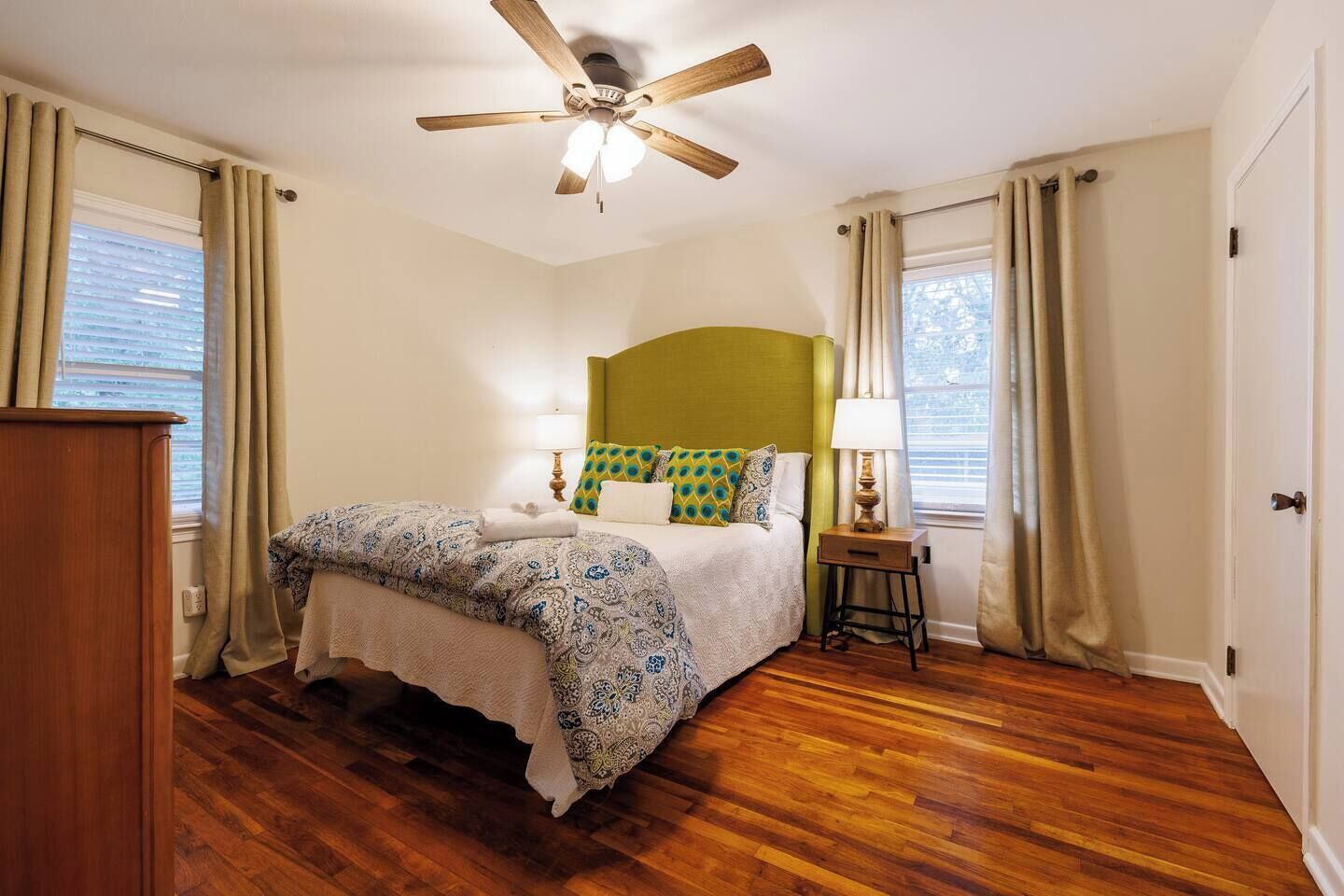 Une chambre avec un plancher en bois, un lit avec une literie à motifs, une tête de lit verte, une table de chevet avec une lampe et un ventilateur de plafond.