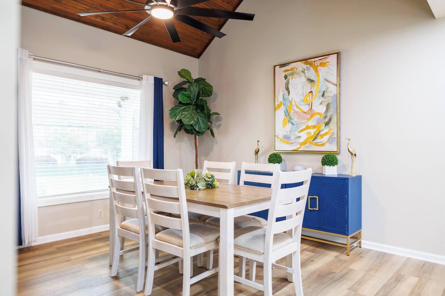 A dining area with a wooden table and white chairs, a blue cabinet, a large plant, and a ceiling fan.