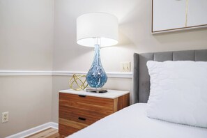 Ein ordentlich bezogenes Bett mit einem weißen Kopfkissen und einem grauen Kopfteil, ein Nachttisch mit einer Lampe und eine wandmontierte Steckdose.
