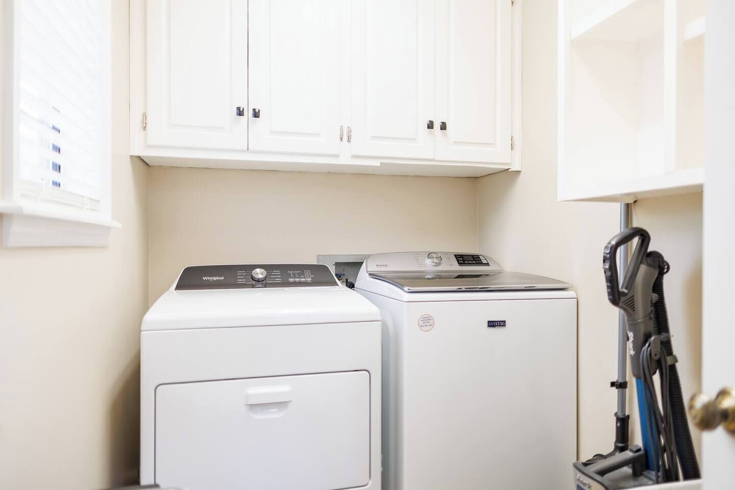 A laundry area with a front-loading washer and dryer, a vacuum cleaner, and white cabinets above.