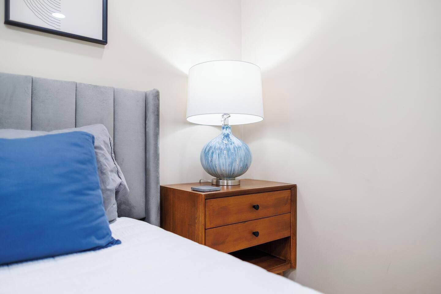 Een opgemaakt bed met een blauw kussen, een houten nachtkastje met een lamp, en een ingelijst beeld aan de muur.