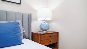 En pænt redt seng med en blå pude, et trænatbord med en lampe og et indrammet billede på væggen.