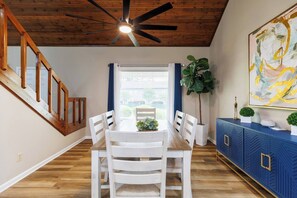 Un coin repas agrémenté d’un ventilateur de plafond en bois, d’une table à manger avec des chaises, d’une fenêtre avec des stores et d’un meuble bleu orné d’objets décoratifs.