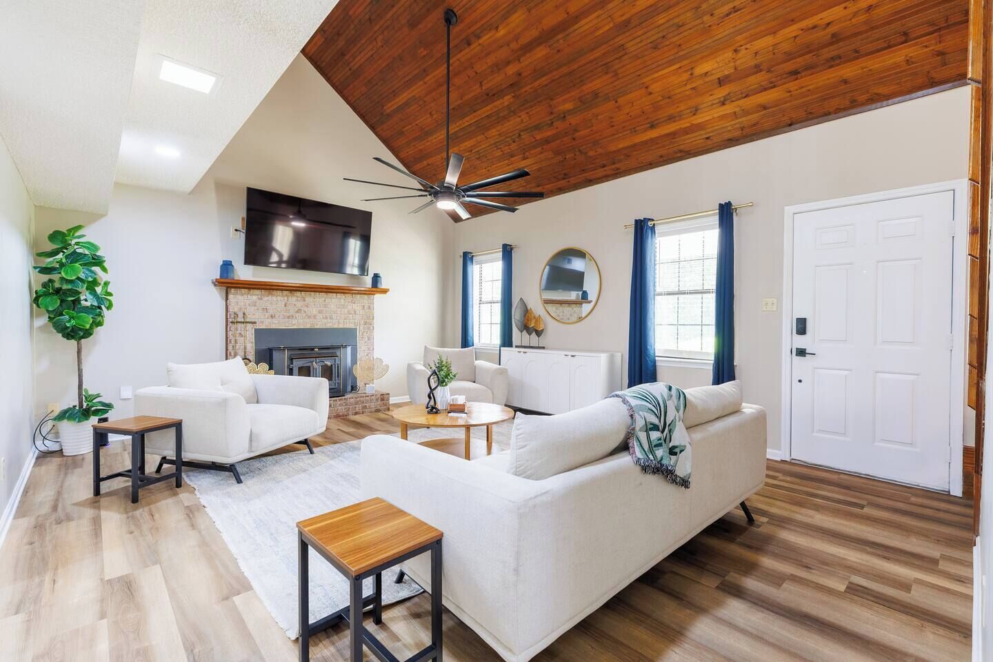 A modern living room with a fireplace, white sofas, a ceiling fan, and a large window with curtains.