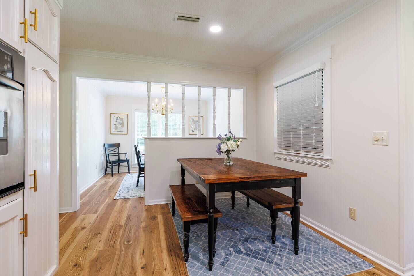 Un comedor con una mesa de madera, bancos y un florero. Se observa una cocina con alacenas blancas y un horno de acero inoxidable. El ambiente cuenta con pisos de madera y un tapete estampado.