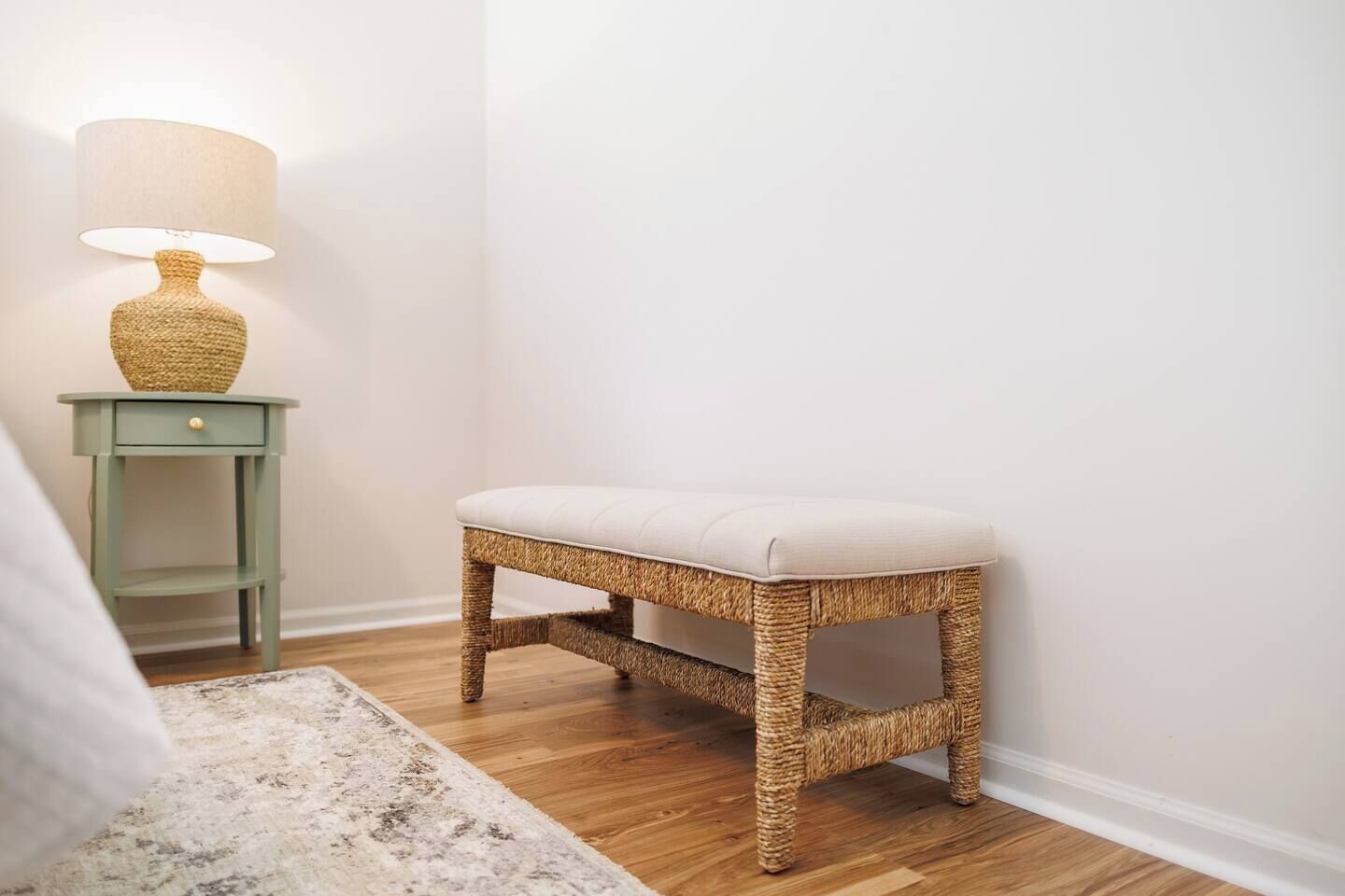 A wicker stool with a cushioned seat, a small green nightstand with a lamp, and a white wall.