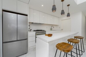A modern kitchen with white cabinetry, a stainless steel refrigerator, a gas stove, and wooden stools. - Heart of Cabo|3BR Oasis|Rooftop Spa|Poolside (Cabo San Lucas)